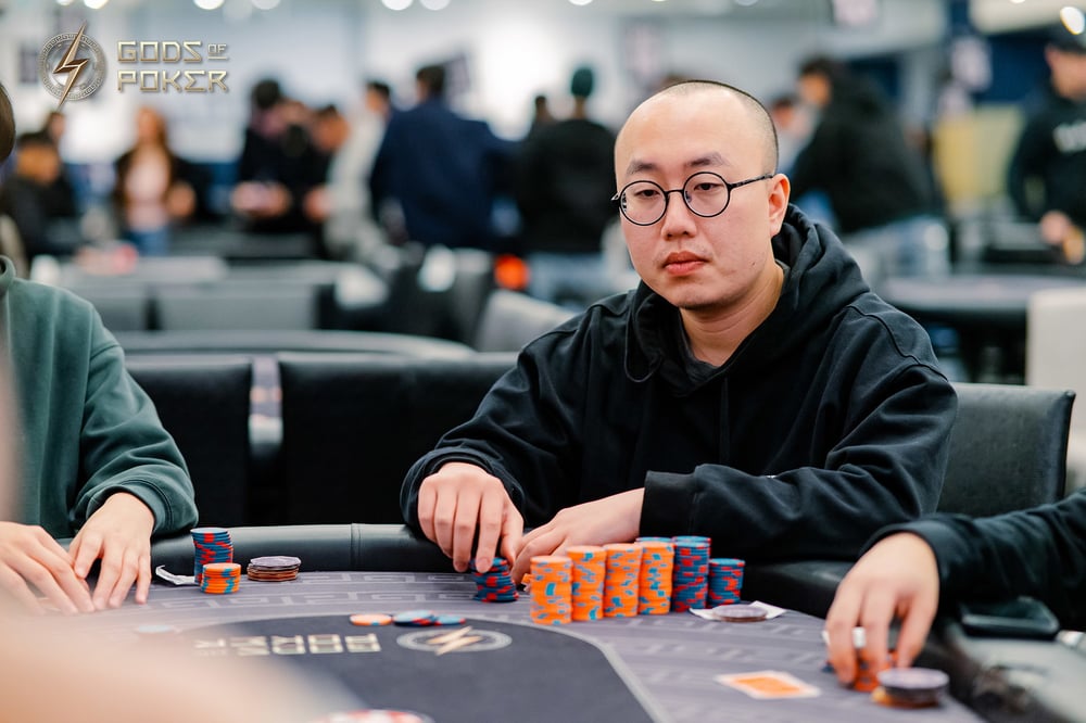 A man with glasses and a black hoodie plays poker, chips stacked on the table.