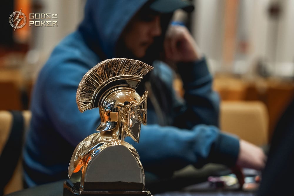 A golden Spartan helmet trophy sits prominently before a blurred poker player in a hoodie.